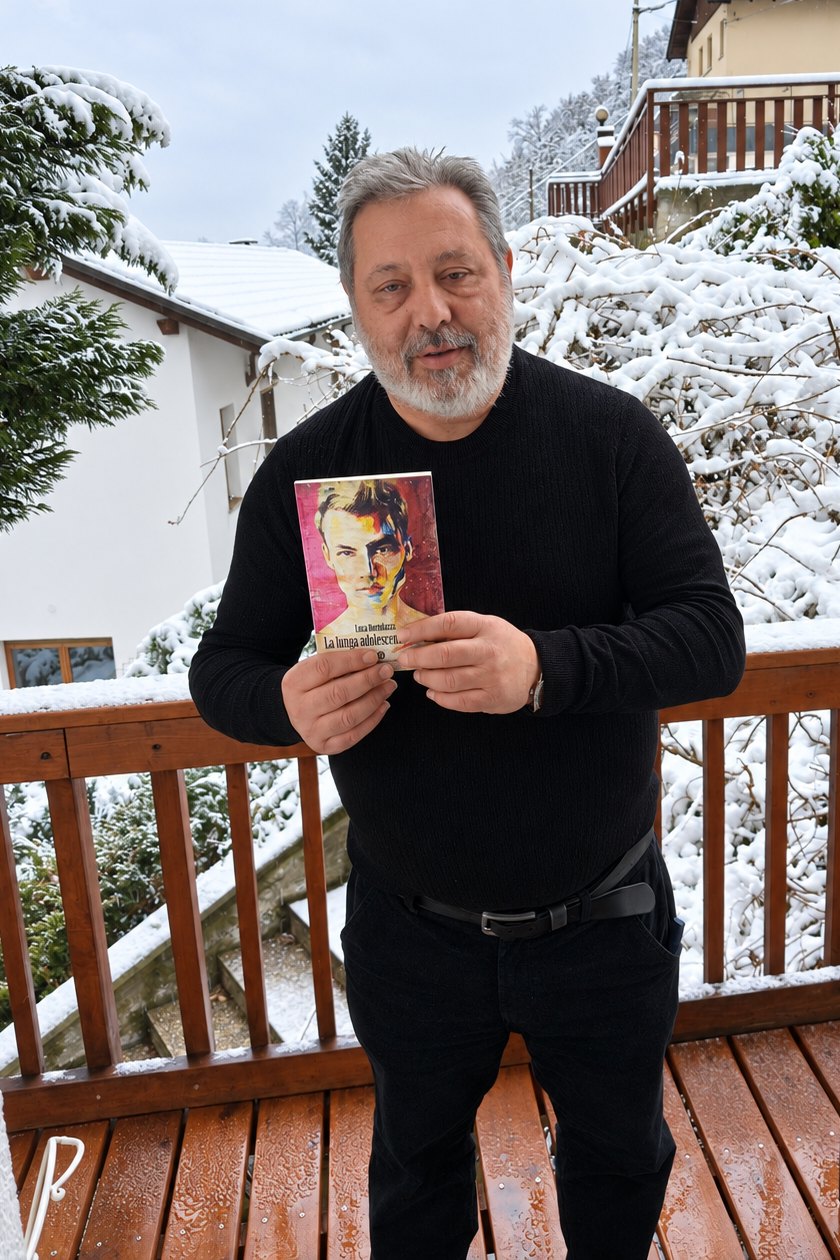 Luca Bortolazzi, scrittore, su un balcone innevato con in mano il suo romanzo La lunga adolescenza di Luca